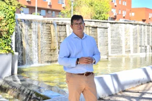 Un hombre con una camisa azul claro y pantalones beige se encuentra de pie frente a una gran fuente al aire libre de la que brota agua en cascada, con edificios de ladrillo rojo al fondo, como si se detuviera un momento en medio de su día para admirar la escena, de forma muy parecida a alguien que revisa un borrador automático antes de ultimar sus planes.