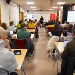 Aula con una veintena de personas asistentes a una formación y una docente exponiendo ante las pantallas de proyección, en Rivas Vaciamadrid