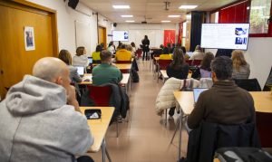 Aula con una veintena de personas asistentes a una formación y una docente exponiendo ante las pantallas de proyección, en Rivas Vaciamadrid