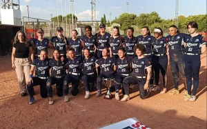 Un equipo femenino de sóftbol, con uniformes azul marino a juego y la inscripción «Riders» en el pecho, posa al aire libre en un campo de tierra. Están reunidas alrededor de un trofeo, algunas arrodilladas y otras de pie, con una mujer de pie en el extremo izquierdo. En representación del CBS Rivas, su unidad y orgullo se hacen evidentes mientras celebran juntas su logro.