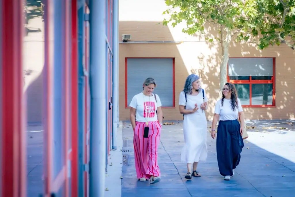 Tres mujeres caminan juntas frente a un edificio con detalles en rojo y azul, disfrutando del animado ambiente de la «vuelta al cole» en Rivas. Una lleva pantalones a rayas rosas, otra lleva un vestido blanco y la tercera lleva un top blanco con una falda oscura. Parecen estar hablando y sonriendo.