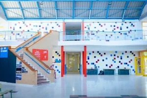 Un luminoso pasillo escolar con azulejos cuadrados de colores en las paredes, una escalera a la izquierda, columnas de soporte rojas y una barandilla en la segunda planta sobre la entrada del pasillo central. El espacio está vacío y bien iluminado por la luz natural, lo que capta a la perfección el animado espíritu de la vuelta al cole en Rivas.