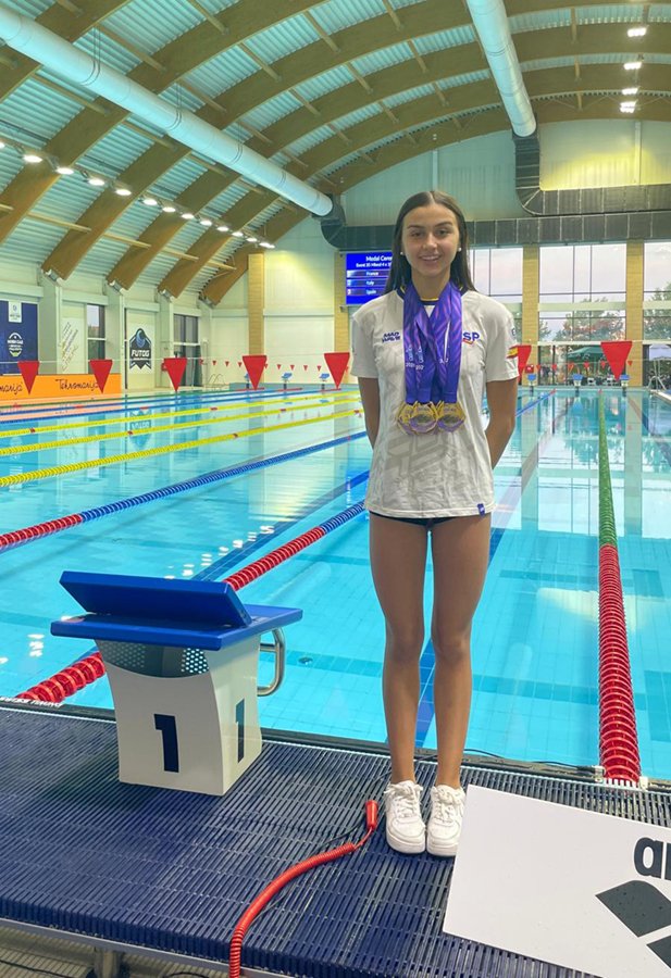 Carlota Rodríguez junto al poyete de la calle 1 de una piscina cubierta, con camiseta de la selección española y tres medallas de oro al cuello