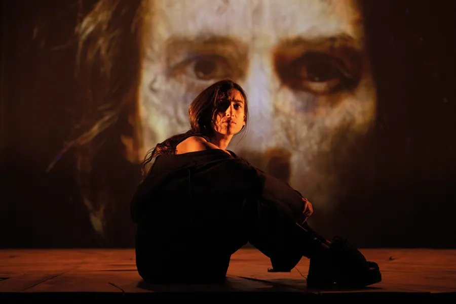Una persona está sentada en un escenario con poca luz, con las rodillas recogidas, y una gran imagen proyectada de un rostro domina el fondo a sus espaldas: una escena impactante que captura el espíritu contemplativo del otoño teatral en Pilar Bardem.