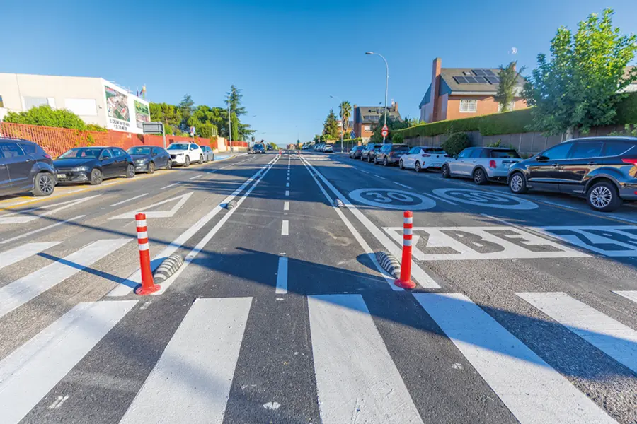 Una amplia calle urbana con marcas viales bien definidas, pasos de peatones blancos y bolardos en el centro. Hay coches aparcados a ambos lados, y edificios y árboles bordean la calle bajo un cielo azul. Un carril bici exclusivo, el «Rivas», discurre junto a la calzada, lo que ofrece a los ciclistas una ruta segura a través del paisaje urbano.