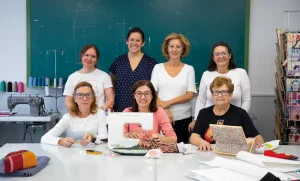 Siete mujeres posan en un aula de costura de la Universidad Popular. Tres están sentadas en una mesa con máquinas de coser y telas, mientras que otras cuatro están de pie detrás de ellas. Al fondo se ven una pizarra, materiales de costura y patrones.