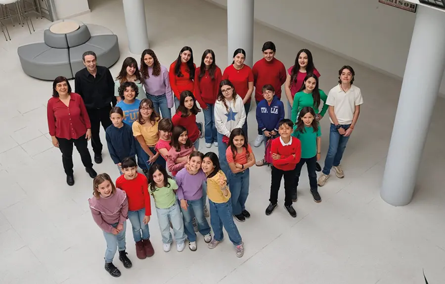 Un grupo de niños y dos adultos están de pie en el interior, sobre un suelo blanco, dispuestos en un semicírculo poco cerrado, mirando hacia la cámara. Los niños, que parecen formar parte de los coros de la Escuela Municipal de Música, llevan ropa de colores vivos; los adultos visten con colores más oscuros. Se ven columnas y mesas blancas.
