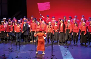Un coro de adultos de los coros de la Escuela Municipal de Música, vestidos de rojo y negro, canta en el escenario bajo la dirección de un director. Uno de los miembros del coro se encuentra de pie detrás del grupo, sosteniendo una bandera roja lisa. El fondo es rojo y hay micrófonos colocados delante del grupo.