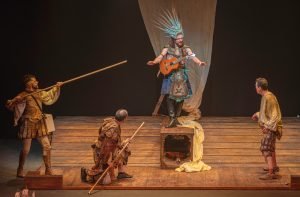 Cuatro actores sobre el escenario en una escena de «La desconquista»: uno con un tocado de plumas y guitarra, otros dos con lanzas y otro con ropas raídas