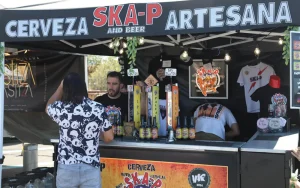 Un hombre se encuentra en un puesto donde se vende cerveza artesanal con el nombre de «Cerveza Ska-P Artesana» en un animado festival celebrado en Rivas. Detrás del mostrador hay dos empleados con grifos de cerveza, botellas y productos con el logotipo de Ska-P, lo que pone de manifiesto su pasión por la cerveza artesanal.
