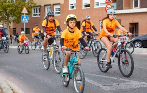 Un grupo de ciclistas, entre los que se encuentran niños y adultos con camisetas naranjas y cascos, pedalea por una calle de la ciudad durante la Fiesta de la Bici en Rivas. En el fondo se ven edificios y señales de tráfico mientras los participantes disfrutan de una de las populares rutas familiares organizadas para el evento.
