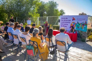Un grupo de personas está sentado al aire libre, de cara a un orador que se encuentra en un atril. Junto a ellas hay una gran pancarta en la que se lee: «Inicio de las obras de la cubierta del metro de Rivas», que destaca el proyecto de la Línea 9B del metro. Dos personas están sentadas en una mesa roja al frente. Al fondo se ven árboles y una valla.