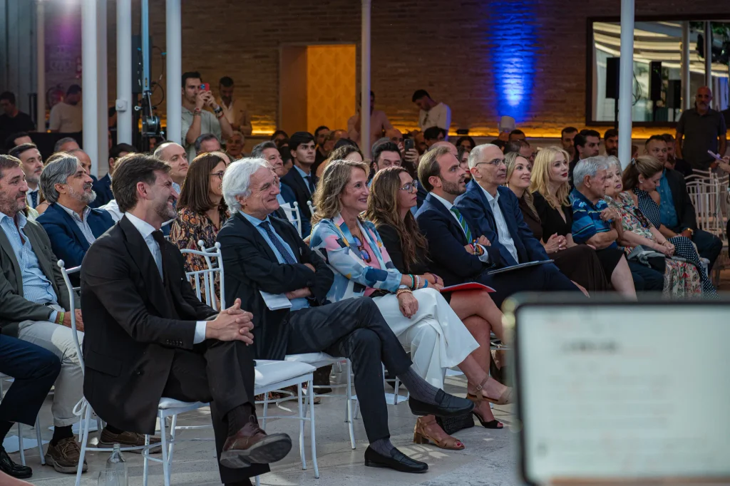Un grupo de adultos, vestidos con ropa formal o de negocios, están sentados en sillas blancas en un acto celebrado en un recinto cerrado, escuchando con atención una presentación durante los Premios Empresariales de ASEARCO. El escenario presenta una decoración moderna, con luces azules que iluminan el fondo.