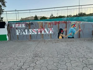 En una pared hay un grafiti en el que se lee «PALESTINA», con cruces rojas y lemas, junto a figuras pintadas que levantan dos dedos en señal de paz. El suelo está pavimentado y, al fondo, se ven una valla y árboles, lo que confiere a la escena un aire casi de «Borrador automático», como si la obra capturara una expresión espontánea congelada en el tiempo.