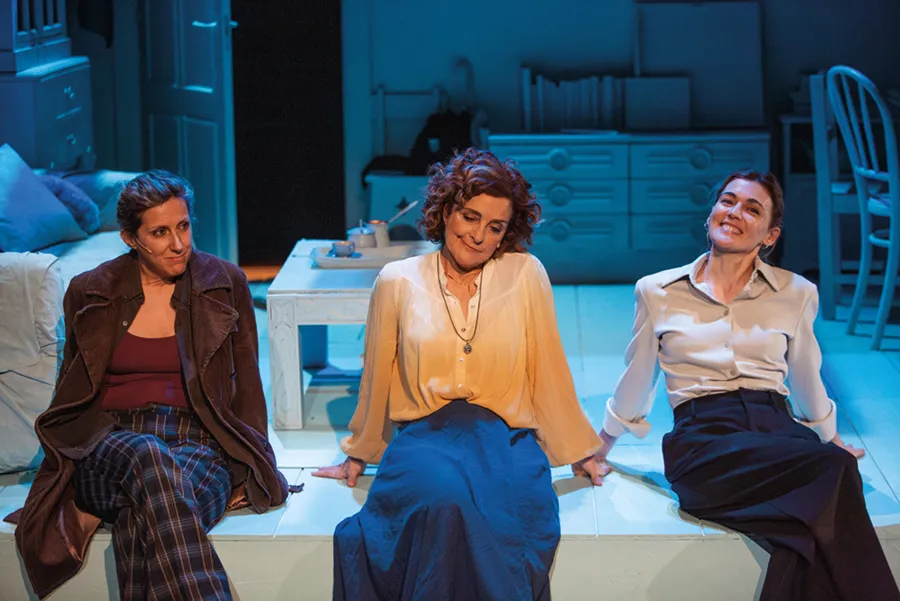 Tres mujeres están sentadas en una plataforma blanca en una sala con iluminación azul durante el Otoño Teatral Pilar Bardem. Llevan blusas y faldas o pantalones. Al fondo se ven muebles, entre ellos una cama y una mesa. Las tres mujeres miran hacia delante, cada una con una expresión diferente.