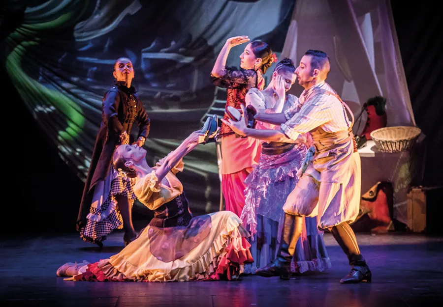 Seis bailarines con trajes de época actúan en el escenario del Otoño Teatral Pilar Bardem, adoptando poses dramáticas con gestos expresivos. El fondo es colorido y abstracto, lo que sugiere una vibrante representación teatral o de danza.
