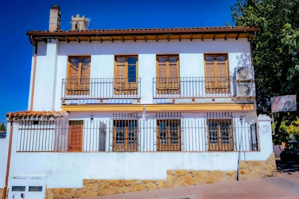 En Rivas se alza majestuoso un edificio blanco de dos plantas con contraventanas de madera, balcones en la planta superior y un zócalo de piedra. Conocida como «La Posada del Alcalde de Rivas», esta encantadora construcción cuenta con ventanas en la planta baja protegidas por rejas metálicas y se alza bajo un cielo azul y despejado tras su cuidadosa rehabilitación.