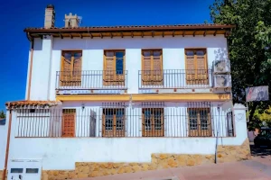 En Rivas se alza majestuoso un edificio blanco de dos plantas con contraventanas de madera, balcones en la planta superior y un zócalo de piedra. Conocida como «La Posada del Alcalde de Rivas», esta encantadora construcción cuenta con ventanas en la planta baja protegidas por rejas metálicas y se alza bajo un cielo azul y despejado tras su cuidadosa rehabilitación.