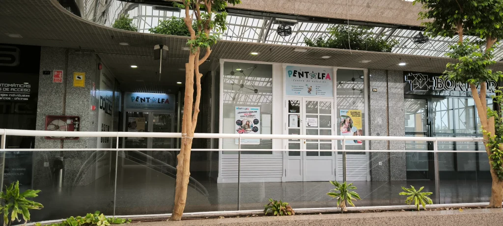 El interior de un edificio moderno con puertas y ventanas de cristal. El letrero situado sobre la entrada dice «PENTALFA». Se ven plantas tanto en el interior como en el exterior, y en las ventanas y puertas hay carteles sobre clases de refuerzo escolar, lo que refleja el compromiso del edificio con el apoyo a las familias locales de Rivas.