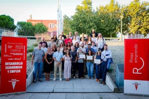 Un grupo de unas 35 personas posa al aire libre en unas escaleras; algunas sostienen certificados que reconocen sus proyectos o empresas. A ambos lados, dos pancartas rojas con los lemas «Rivas Emprende» y «Desarrollo Económico y Empleo» resaltan el espíritu de los Premios de Rivas. Al fondo se ven árboles y un edificio de ladrillo.