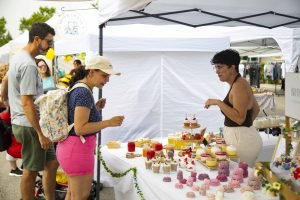 Personas contemplando la mesa de un puesto de repostería artesanal, con dulces y tarros, bajo una carpa blanca en un mercado al aire libre