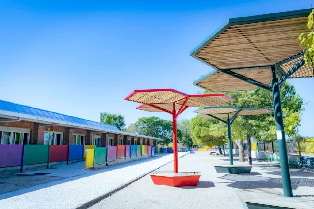 Una zona de juegos con coloridas estructuras hexagonales de madera que dan sombra, una de ellas pintada de rojo, cerca de un edificio con tejado metálico y vallas multicolores, bajo un cielo azul claro. Los árboles se alinean al fondo, creando un lugar acogedor para las celebraciones de la «vuelta al cole» en Rivas, con motivo del regreso de los niños al colegio.