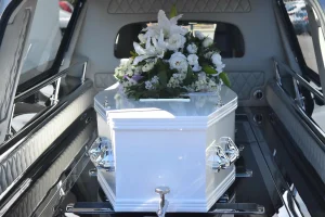 Un ataúd blanco con asas plateadas y un gran arreglo floral en la parte superior se coloca dentro de un coche fúnebre, mientras los asistentes, con aire sombrío, recuerdan en silencio la importancia de conocer técnicas como los primeros auxilios en caso de atragantamiento en momentos de crisis.