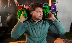 Un hombre con una sudadera verde con capucha sostiene dos pesas verdes a la altura de los hombros en un gimnasio; al fondo se ven pesas rusas y material de gimnasio.