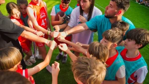 Un grupo de niños y dos adultos están de pie formando un círculo sobre la hierba, estirando los brazos hacia el centro para chocar los puños entre ellos. Los niños llevan petos deportivos y hace sol, lo que crea una escena perfecta para la diversión de un campamento de verano inspirado en los Miami Dolphins.
