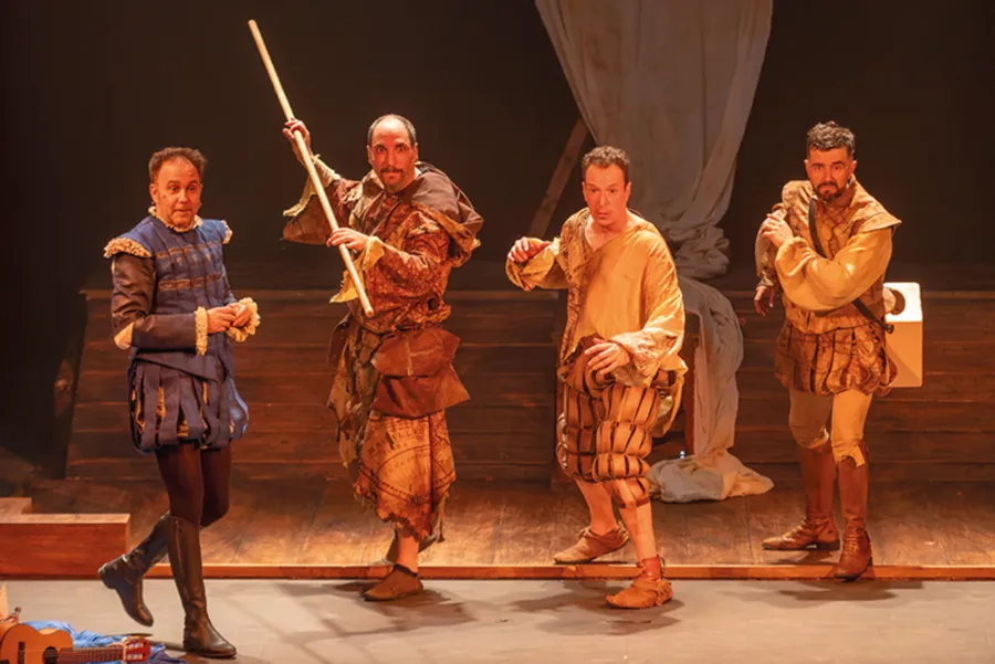 Cuatro actores vestidos con trajes renacentistas actúan en un escenario de madera en tonos beige y marrón, adoptando poses dramáticas que recuerdan a un «otoño teatral» de Pilar Bardem. Al fondo cuelga un gran telón drapeado y se ven los elementos escenográficos.