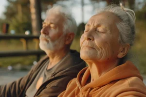 Un hombre y una mujer de edad avanzada están sentados al aire libre con los ojos cerrados, de cara a la luz del sol. Ambos parecen relajados; la mujer lleva una sudadera con capucha naranja. Mientras disfrutan juntos de este momento de paz, el entorno tranquilo refleja sutilmente temas relacionados con el envejecimiento molecular. En el fondo se ven borrosos unos árboles y una estructura de madera.