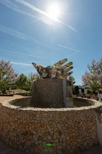 Una fuente de piedra en el Jardín de la Paz de Rivas, con una escultura central que representa un pájaro con forma de mano que refleja la luz bajo un cielo soleado.
