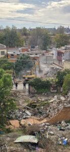 Excavadora derribando una infravivienda en la Cañada Real, con escombros en primer plano y agentes de seguridad