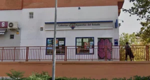 Una oficina de Loterías y Apuestas del Estado en Rivas, con una barandilla metálica roja en la fachada y una persona que pasa junto al edificio.