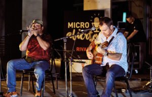 Dos músicos tocan la armónica y la guitarra en el escenario del Micro Abierto de Rivas, con el cartel del programa al fondo