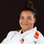 Nisrin Bousbaa, vestida con un uniforme blanco de judo y un cinturón negro, sonríe con los brazos cruzados delante del logotipo de la Federación Internacional de Judo.