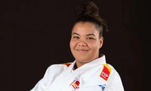 Nisrin Bousbaa, vestida con un uniforme blanco de judo y un cinturón negro, sonríe con los brazos cruzados delante del logotipo de la Federación Internacional de Judo.
