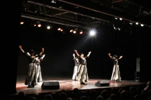 Cinco artistas, con camisetas negras y faldas largas estampadas, bailan en un escenario bajo brillantes focos para promover la solidaridad a través del arte.