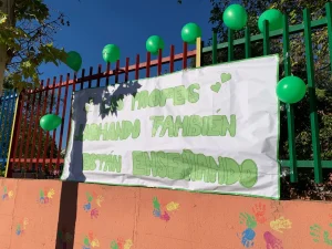 En una valla hay una pancarta en la que se lee Los profes luchando también están enseñando, rodeada de globos verdes que representan la marea verde y de coloridas huellas de manos en la pared que hay debajo.