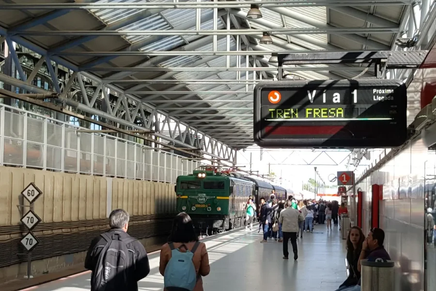 La gente camina por un andén cubierto mientras se acerca un tren verde, lo que evoca el encanto de los trenes históricos de la Comunidad de Madrid. Un letrero electrónico indica: «Vía 1 TRENE FRESA». El andén cuenta con vigas metálicas, paneles de cristal y diversos letreros visibles, todo ello bañado por la suave luz del otoño.