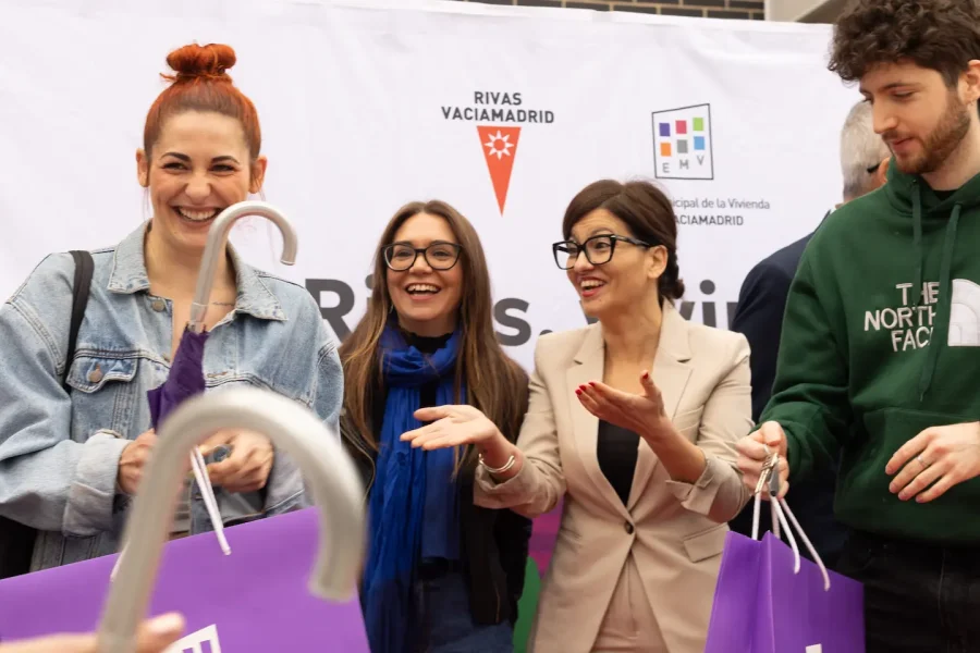 Cuatro personas se sitúan frente a una pancarta de Rivas, sonrientes y con bolsas moradas en las manos. Dos mujeres en el centro gesticulan con las manos, mientras un hombre con sudadera verde mira un objeto relacionado con alquiler para jóvenes. En primer plano se ve el mango de un paraguas.