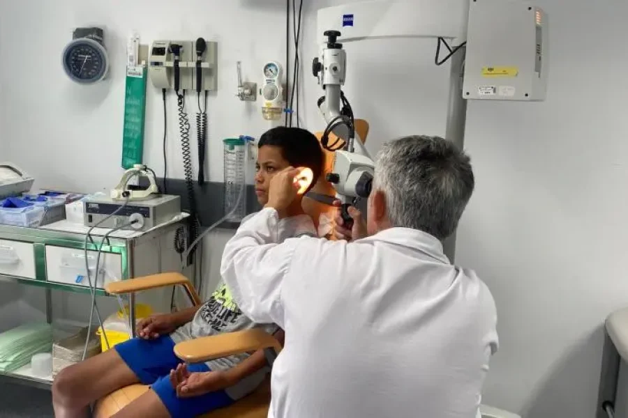 Un médico examina el oído de un niño con un instrumento médico en la consulta de una clínica, lo que pone de relieve la importancia de la salud de los menores. En el fondo se ven equipos médicos, material sanitario y un reloj de pared.