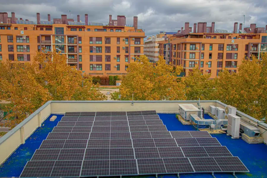 Una azotea de Rivas presenta un gran conjunto de paneles solares de autoconsumo junto a unidades de climatización, con árboles otoñales y varios edificios residenciales naranjas de fondo bajo un cielo nublado, posiblemente parte de iniciativas de talleres municipales.