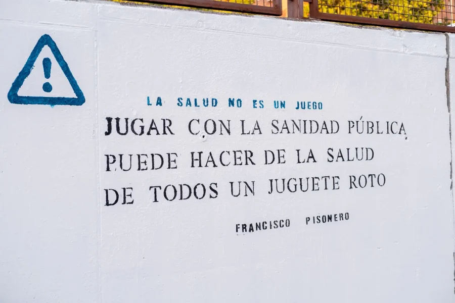 Una pared blanca en Rivas con un triángulo de advertencia pintado y un texto en español que dice: La salud no es un juego. Jugar con la sanidad pública puede hacer de la salud de todos un juguete roto. Francisco Pisonero, poesías.