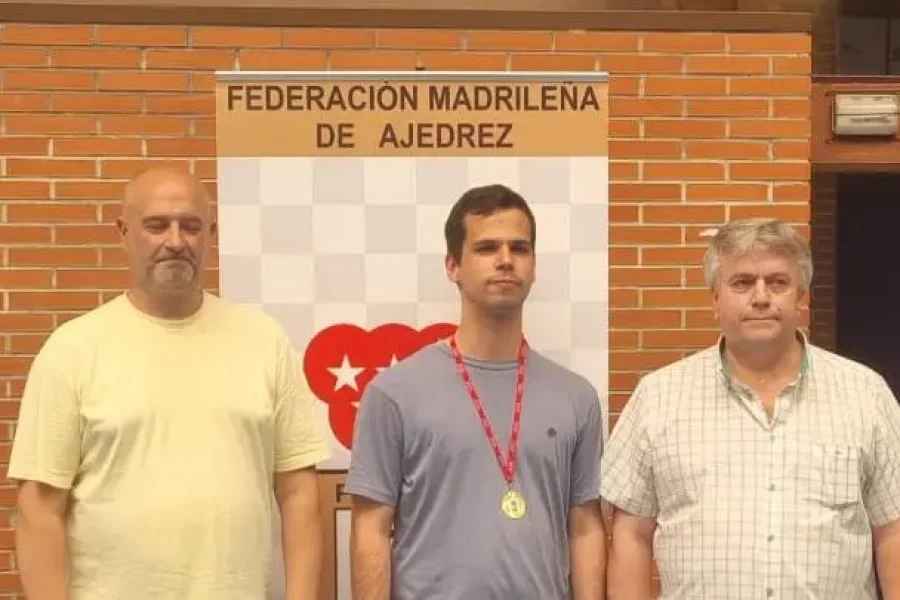 Tres hombres posan delante de un cartel de la Federación Madrileña de Ajedrez. El hombre del centro lleva una medalla de oro y una camiseta gris, mientras que los otros dos se sitúan a ambos lados de él, todos mirando a la cámara.
