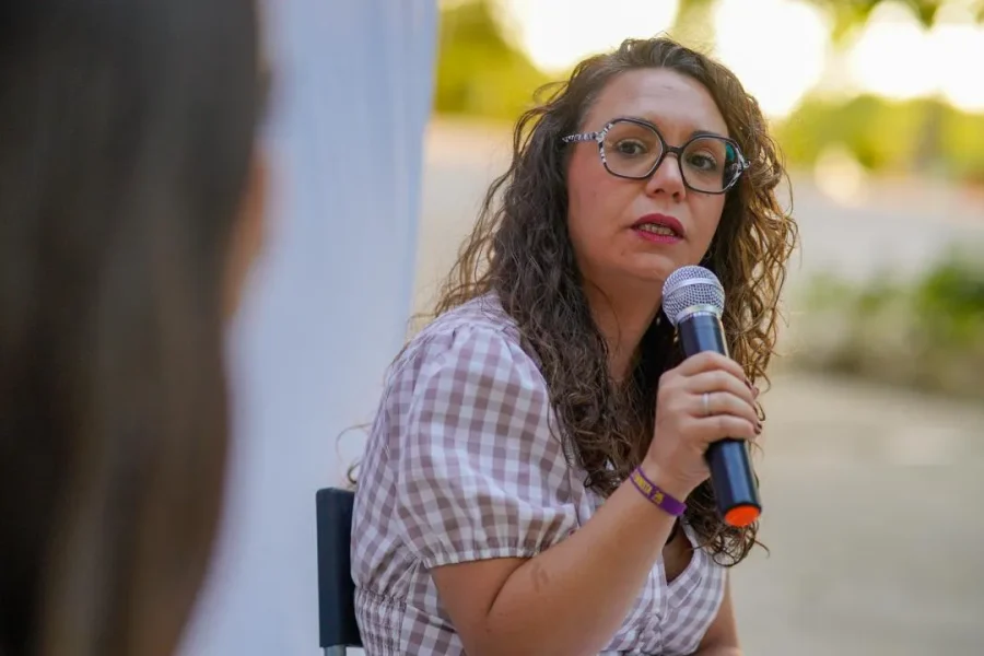 Aída Castillejo, una mujer de pelo rizado y con gafas, sostiene un micrófono y habla mientras está sentada al aire libre. Lleva una blusa estampada y mira hacia otra persona cuya espalda se ve en primer plano.