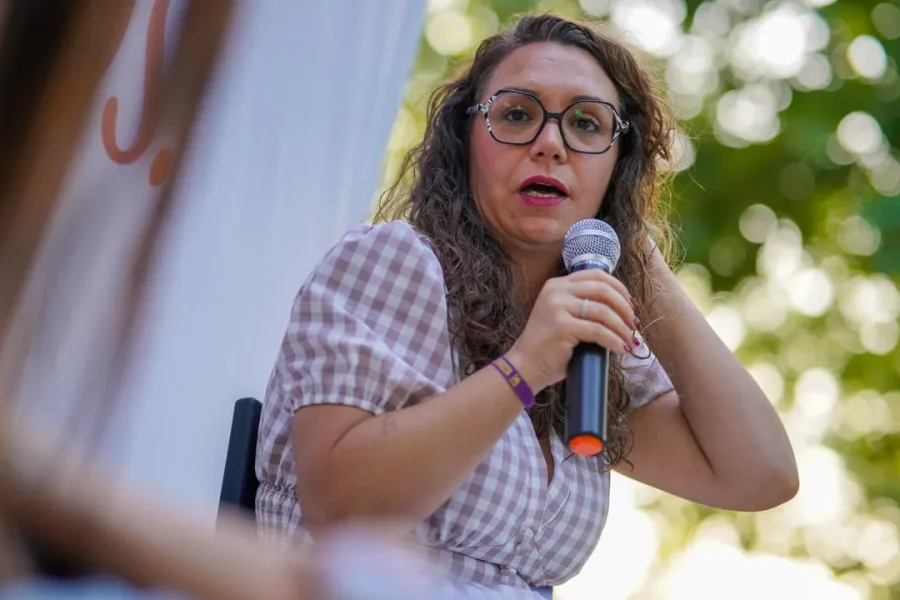 Aída Castillejo, vestida con un vestido a cuadros, está sentada al aire libre en Rivas, con el pelo largo y rizado y gafas, sosteniendo un micrófono mientras habla —con una mano tocándose el pelo— ante un fondo de árboles verdes difuminados.