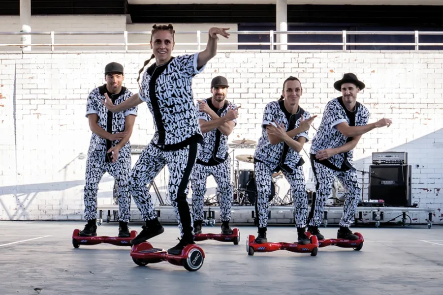 Cinco personas vestidas con trajes a juego en blanco y negro realizan una coreografía sobre hoverboards frente a una pared de ladrillo blanco, con baterías y altavoces visibles al fondo: una escena llena de energía del Festival de Cultura en la Calle 2026.