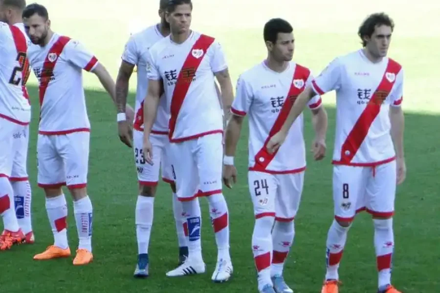 Un grupo de jugadores de fútbol con uniformes blancos con una franja diagonal roja, que recuerdan al Abonos Rayo Vallecano, de pie sobre un campo de hierba. En primer plano se ven cuatro jugadores, algunos mirando a los lados y otros con los brazos a los lados.