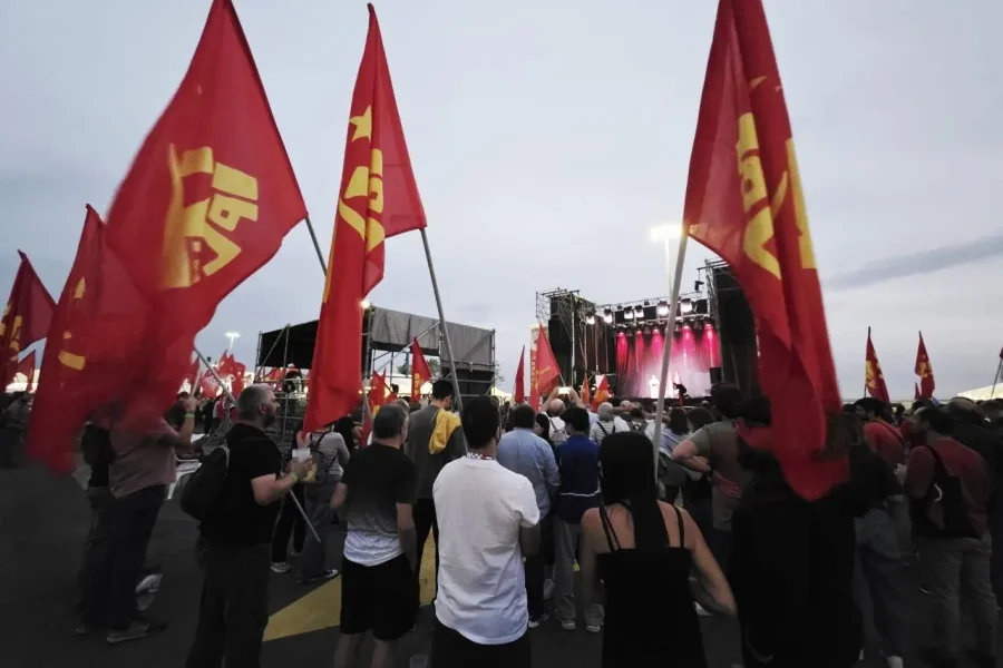 Una multitud se reúne al aire libre en Rivas, ante un escenario en las Fiestas del PCE. Banderas rojas con símbolos y textos amarillos ondean mientras luces rosas iluminan la escena, y la gente observa la actuación a pesar del cielo nublado.